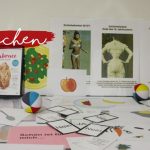 Auf einem Tisch liegen Unterrichtsmaterialien aus, darunter Karten, Broschüren, bunte Strandbälle und Poster mit Abbildungen von Menschen und Gegenständen. In der linken oberen Ecke steht in roter Schreibschrift der Satz "Fit machen".