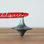 Ein Kreisel balanciert auf einer Holzfläche. Darüber steht in weißer Handschrift auf einem roten Farbstreifen das deutsche Wort sensibilisieren. Der Hintergrund ist einfarbig und hell gefärbt.