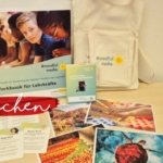 Auf einem hellen Holztisch sind ein Arbeitsbuch, eine Tragetasche, Karten und Postkarten ausgestellt. Die Materialien stammen aus dem Projekt #mindfulmedia, das sich an Lehrerinnen und Lehrer richtet, mit rot überlagert steht "fit machen" unten links.
