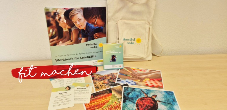 Auf einem hellen Holztisch sind ein Arbeitsbuch, eine Tragetasche, Karten und Postkarten ausgestellt. Die Materialien stammen aus dem Projekt #mindfulmedia, das sich an Lehrerinnen und Lehrer richtet, mit rot überlagert steht "fit machen" unten links.