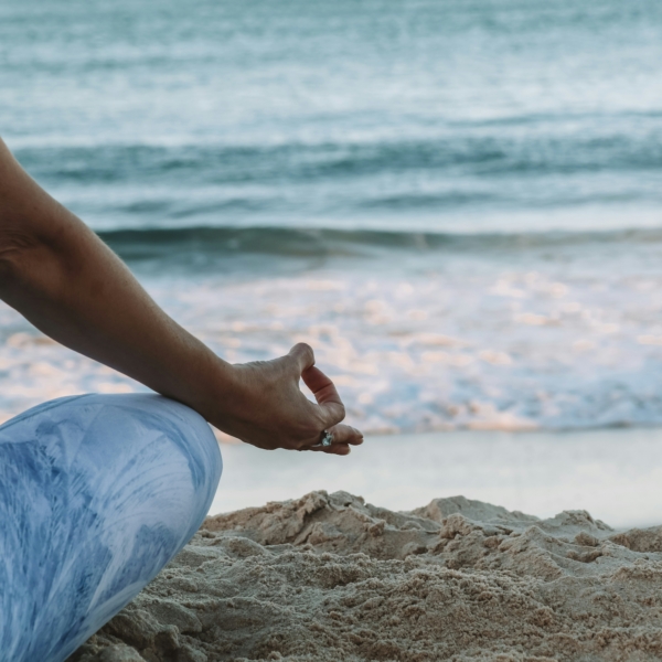 Zu sehen ist der Anschnitt einer Frau von hinten, sitzend im Schneidersitz mit typischer Yoga-Fingerstellung. Sie sind im Sand, vor ihr das Meer.