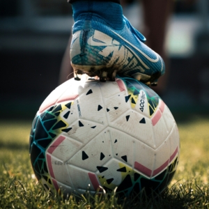 Ein Fußball liegt auf dem Rasen, darauf steht ein Fuß in Fußballschuhen.
