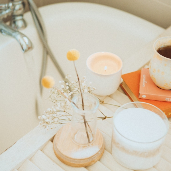 Man sieht einen Ausschnitt einer Badewanne, mit Blümchen , Kerze und einer Tasse Kaffee am Badewannenrand. Das Licht ist gemütlich in Beige- und Gelbtönen gehalten.